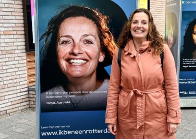 Een vrouw met een oranje jas en bruin haar staat voor een fotokubus waar Faces of Rotterdam op staat met haar foto in de vorm van een R.
