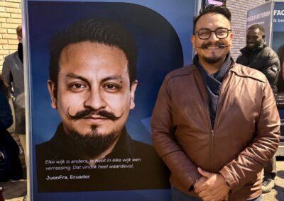 Een getinte man met bril en snor in een bruine jas staat voor een fotokubus waar Faces of Rotterdam op staat met zijn foto in de vorm van een R.