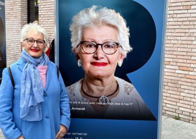 Een vrouw met blauwe jas, wit haar en rode lippen staat voor een fotokubus waar Faces of Rotterdam op staat met haar foto in de vorm van een R.