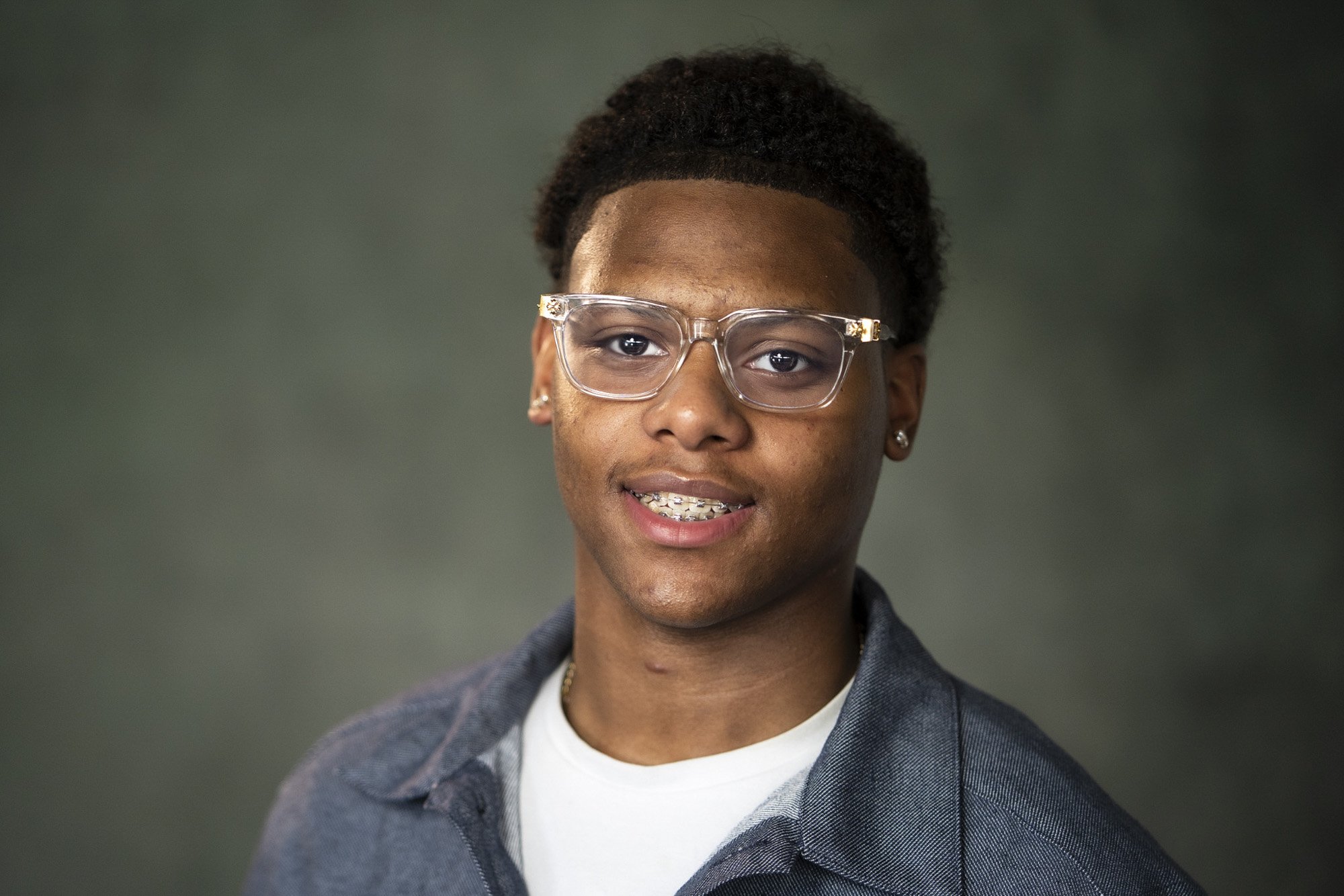Je ziet een jonge ekleurde jongen met een doorzichtige bril en zwart haar voor een groene achtergrond naar de camera kijken